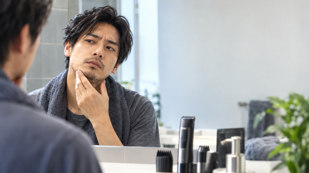 鏡でヒゲの状態を確認する男性の前に、ヒゲトリマー本体と複数のコーム型アタッチメントが並ぶ洗面台