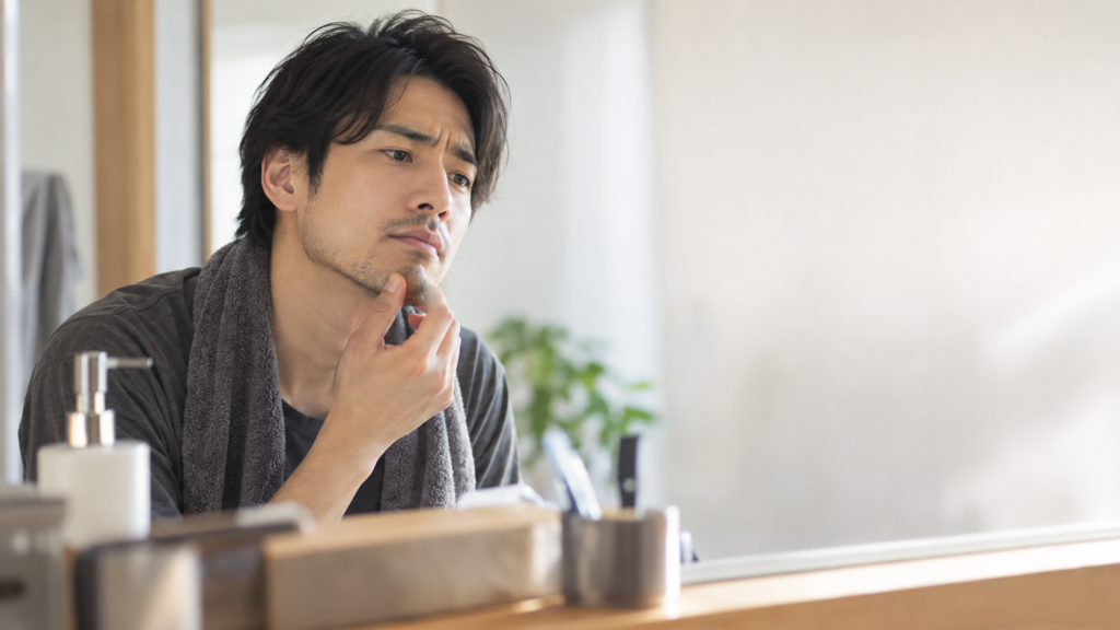洗面台の鏡でヒゲの生え方を確認し、あごに手を添えて長さや整え方を考える男性