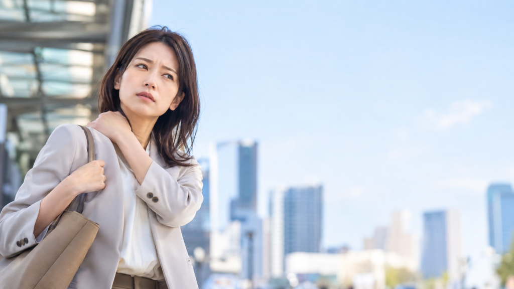 都市部の屋外でトートバッグを肩に掛け、首元に手を添えながら立つ女性