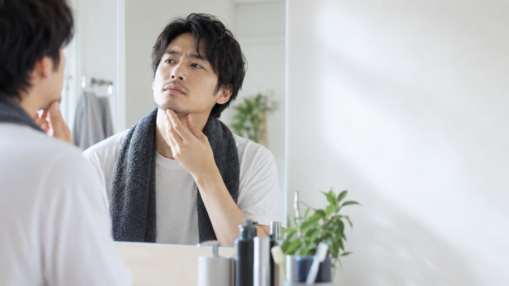明るい洗面所であご下のヒゲを手で確認し、トリミング前の長さを見極める男性