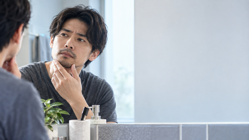 洗面所の鏡越しに口元とあごのヒゲを見ながら、輪郭や伸び具合を確かめる男性と洗面用品