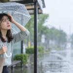 雨のバス停で傘とバッグを持ち、濡れた道路を見ながら立ち止まる女性