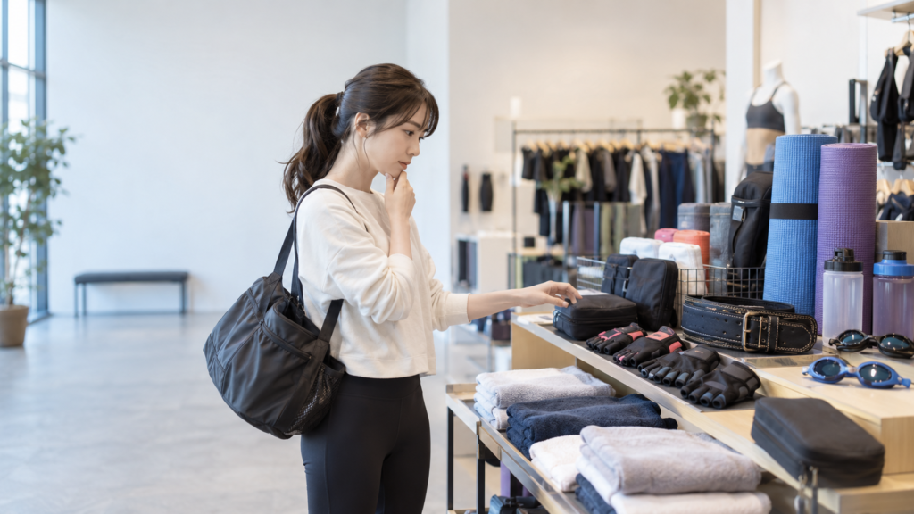 トレーニンググローブやベルト、ヨガマットなどが並ぶ売り場で用品を選ぶ女性